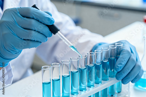 Scientist adding blue liquid to test tubes in laboratory with pipette and protective gloves