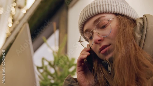 Young white woman on phone in intimate storefront closeup, wearing beanie and glasses with winter jacket, listening intently and expressing concern, soft holiday lights and bokeh create empathetic