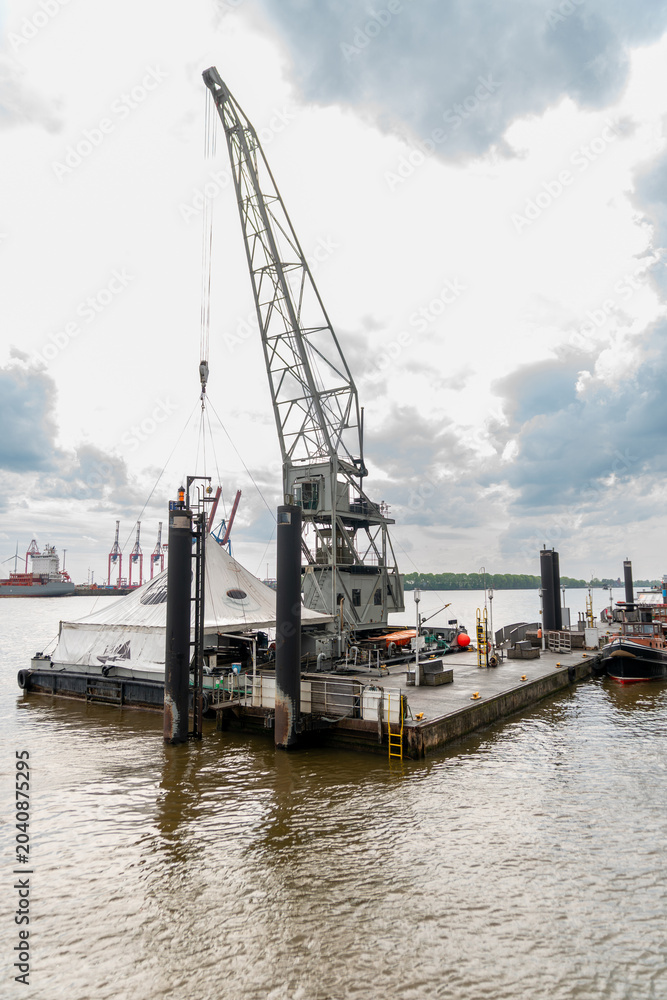 Obraz premium Industrial floating crane platform anchored on a wide harbor river with steel lifting structure, mooring posts, cloudy sky, and maritime port equipment.