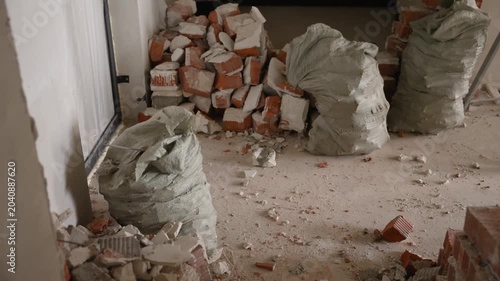 room renovation construction site with stacked bricks and ladder on dusty floor, natural light through door, messy environment with debris and sacks, unfinished walls and renovation materials