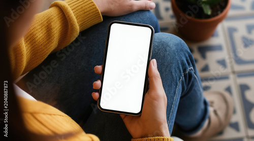 Woman holds blank smartphone screen. Person uses mobile device for social media or online shopping. Female with phone on lap sits near potted plant on tiled floor.