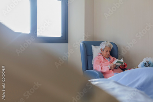 Elderly Woman Reading Book In Nursing Home Chair Promoting Healthy Aging And Senior Well-Being In Healthcare Setting