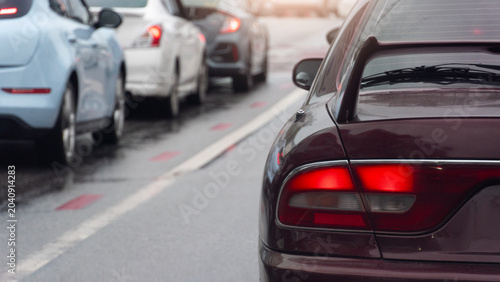 Rear side of a dark red sports car stuck in heavy daytime traffic. Long line of white cars stretches out ahead along the multi-lane city avenue under bright daylight and on a rain soaked road.