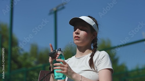 Tired female tennis player in a visor resting and drinking water from a bottle after a match. Athletic woman rehydrating during a break in training