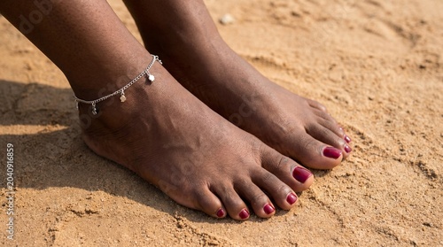 Dark skin feet with burgundy pedicure and silver anklet on sand