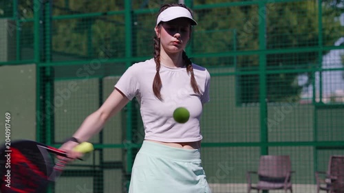Young athletic woman with braids serving a tennis ball with a padel racket. Female player practicing with concentration on a green court