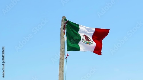 Mexican flag in the wind, blue sky in the background. Wooden pole with mexican state flag in the fresh wind.
