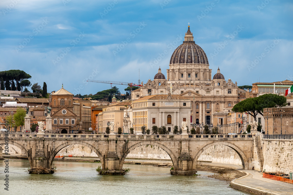 Fototapeta premium View on stone-arched bridge on Tiber river and St Peter's Basilica in Rome, cloudy sky, ochre and terracotta buildings, domes and umbrella pines rise on gently sloping Vatican hill