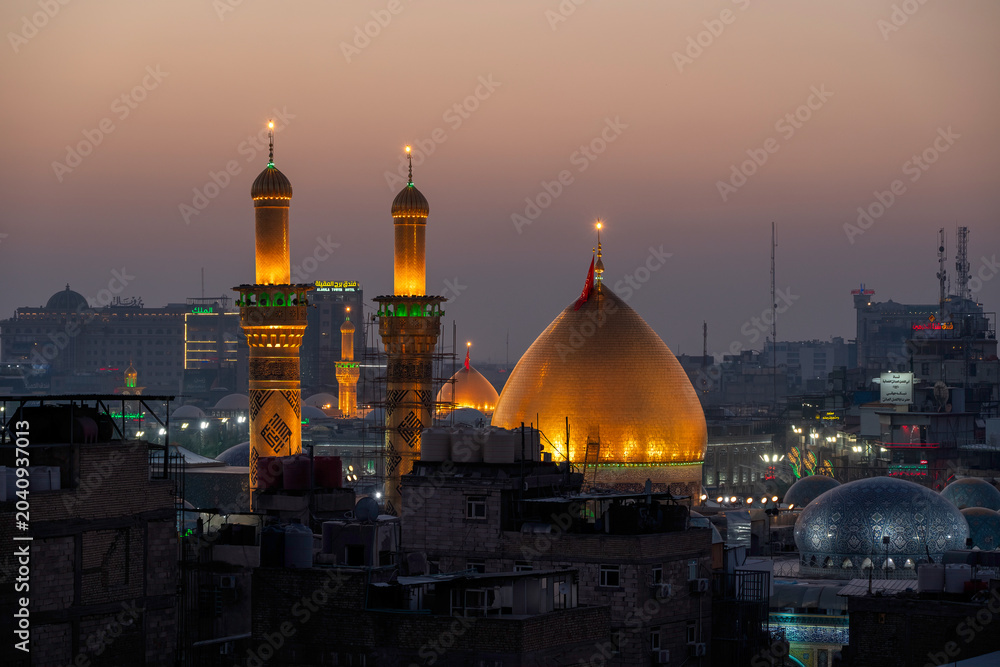 Fototapeta premium Karbala, Iraq - December 28, 2025: Golden dome and illuminated minarets of the Imam Hussain Shrine glowing at dusk in Karbala, Iraq