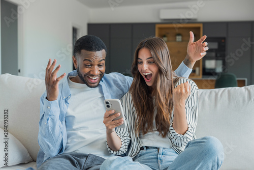 Fotografie Young couple on a couch celebrating together as they react with joy to winning n