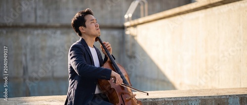Asian cellist playing cello outdoors at concrete structure in warm golden light, elegant male musician performing classical music on instrument, serene and focused creative artist expression.