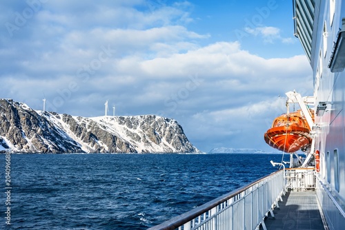 View from a ferry or cruise ship deck sailing in Norway to the snow covered mountains with wind turbines are visible in the distance