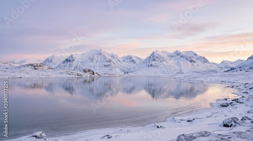 Serene winter landscape of snow-covered arctic mountains perfectly reflecting in a calm icy lake during a pastel pink and purple sunset, ideal for serene travel and nature background concepts.
