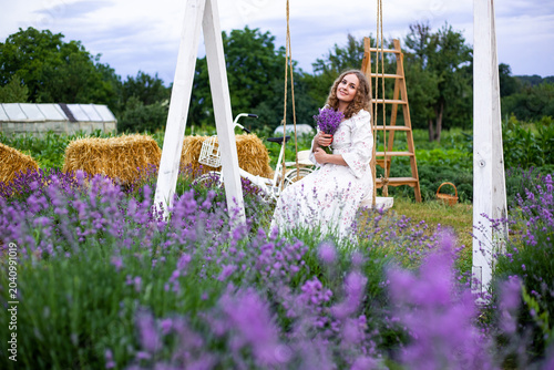 Stylish woman sits on swing in a lavender field in a white dress with a bouquet of lavender in her hands, Provence vibe, summer