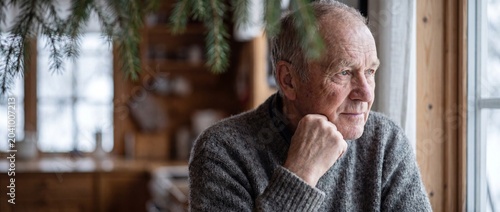 Senior man looking out window in cozy cabin thinking. Elderly thoughtful male in grey sweater contemplating life by glass pane. Pensive mature adult in retirement home feeling lonely and nostalgic.