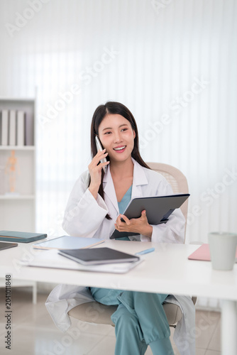 Asian medical doctor talking phone consultation patient holding tablet device healthcare professional white coat modern clinic office workspace telemedicine