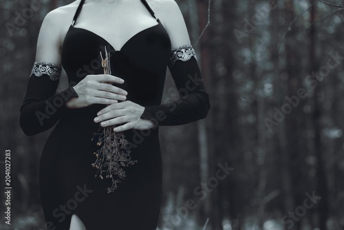 Quadro su tela Faceless witch in gothic black dress and deathly pale skin holding dry plants in hands in dark spooky forest, copy space