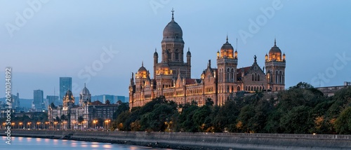 The image showcases the magnificent heritage architecture of Mumbai, illuminated at dusk, presenting a regal facade along the waterfront against a vivid skyline.