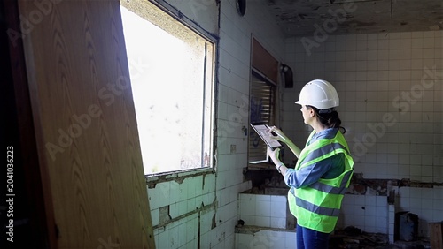 Bild auf Leinwand Woman wearing hard hat and reflective vest documenting structural issues and sur