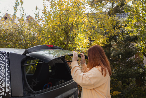 Rear view of a young woman with long hair putting bags into an open car trunk while preparing for a weekend travel journey outside in autumn nature.