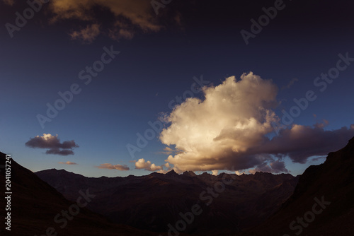 Obraz na płótnie Awesome alpine panorama with peaks silhouettes and sunlit clouds at sunset, Vall