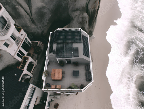 Upward-facing drone shot hovering just below a cantilevered rooftop on a cliffside home, where an installation team in harnesses fits frameless solar modules along a wind-blown edge, ocean waves crash