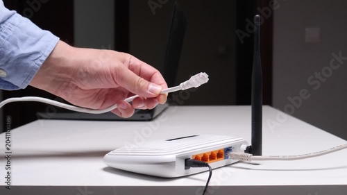 Person's hand connecting a white ethernet patch cord to a wireless internet router on a white table, establishing network connectivity with a laptop in the background