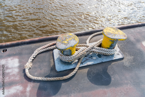 Two yellow steel mooring bollards wrapped with thick rope on a worn boat deck beside rippled harbor water. Useful for maritime transport, docking, nautical equipment, river travel, and industrial wate