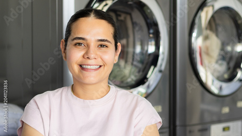 Fotografía Portrait of a smiling young woman standing confidently inside a modern laundromat, with industrial front-loading washing machines in the background