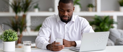 A man with a beard sits at a desk, looking at his smartphone while working on a laptop, surrounded by greenery, reflecting a modern and productive workspace atmosphere.