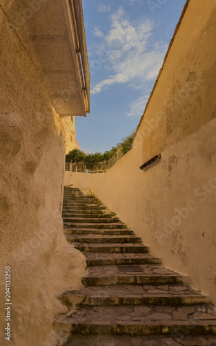 A low-angle vertical shot of an ancient stone staircase winding upwards through a narrow alleyway with textured, sun-drenched walls, perfect for travel, architecture, and lifestyle themes.