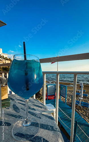 Vibrant blue cocktail on a sunny beach terrace. A vertical close-up shot featuring a bright blue cocktail in a large glass with a straw, placed on a table with a fish-patterned tablecloth.