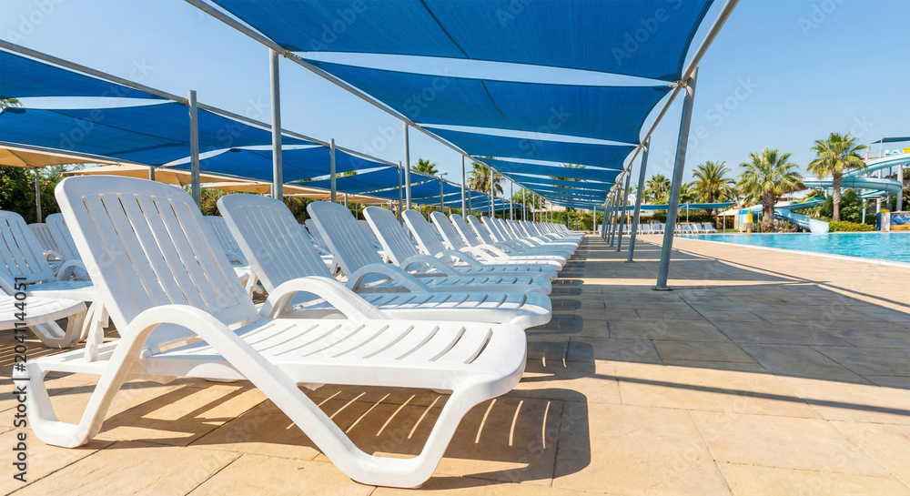 Fototapeta premium A neat row of empty white plastic deck chairs rests in the shade of blue fabric awnings next to a sunny outdoor swimming pool.