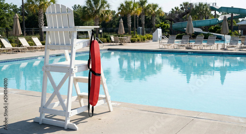 A white wooden lifeguard chair with a red rescue tube hangs by a bright blue outdoor swimming pool on a sunny day.