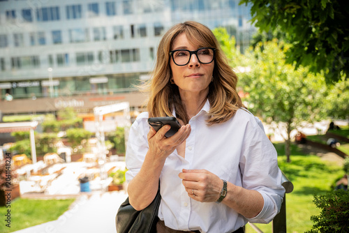 Portrait of a smiling professional woman with eyeglasses holding a smartphone in city park, modern business lifestyle