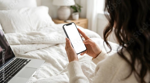 Woman holding smartphone with blank white screen on bed with laptop.