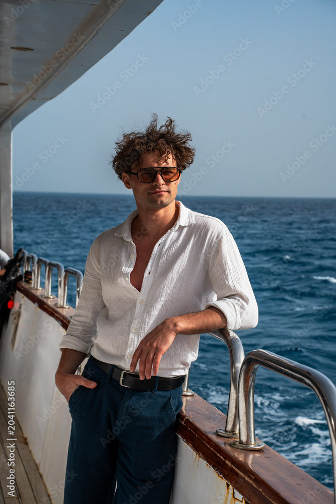 Fototapeta premium Epic tropical cruise traveler man luxury yacht vertical portrait. Cinematic full length view of confident wealthy male explorer on boat deck against blue ocean water and horizon sky. 