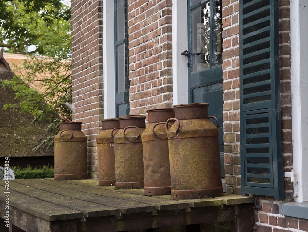 Obraz premium Several vintage rusty metal milk cans standing on a weathered wooden porch near brick walls