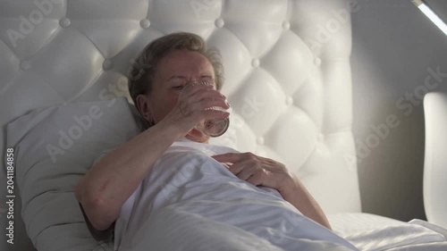 A sick elderly woman drinks water in bed at night