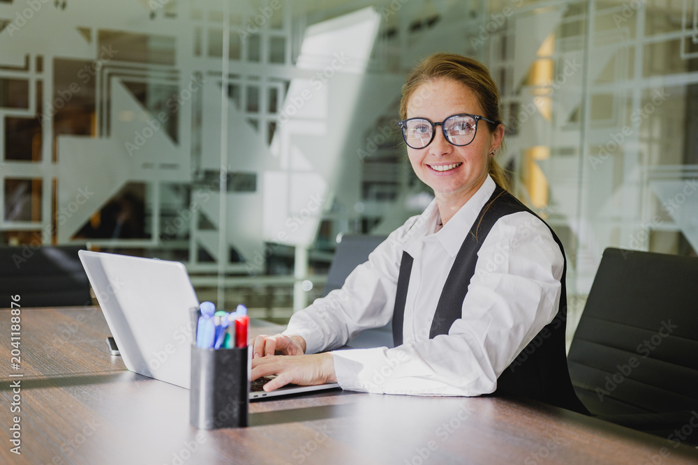Fototapeta premium Professional woman working at her desk in a modern office setting while smiling and typing on a laptop during a bright afternoon