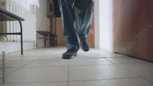 Black paramedic walking down hallway toward door, blue scrubs and black shoes on tile floor, lowangle shot showing bench and radiator, steady footsteps conveying urgency and focus while approaching
