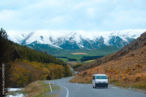 South Island, New Zealand: Roadtrip view of white passenger van driving along winding asphalt highway below a snow covered alpine mountain range.