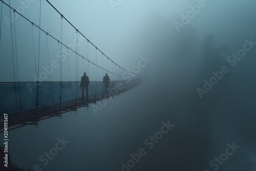 Silhouettes of two people on a long suspension bridge disappearing into dense, blue-toned fog, creating a mysterious and atmospheric scene.