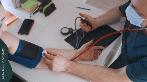Home visit white woman arm measurement, paramedic nurse fits cuff and records systolic and diastolic readings, maskwearing clinician offers calm reassurance, domestic desk with phone and paperwork,
