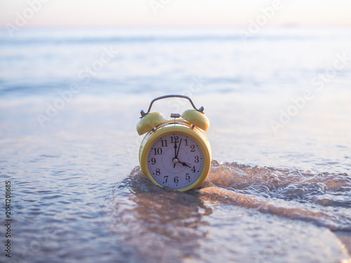 A yellow twin-bell alarm clock sitting on the sand as sea waves wash over it
