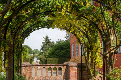 London, UK, Kew garden palace. A scenic view of a garden terrace framed by a metal archway covered with blooming yellow laburnum flowers.  