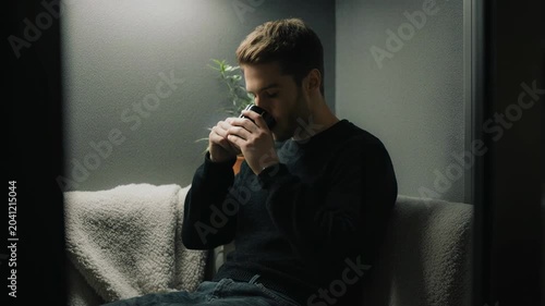 Slow tracking shot of man holding and lifting mug while seated on couch in dim living room during evening