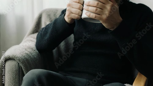 Static close up slow pan of person holding coffee cup and sipping while sitting in armchair in living room with soft natural morning light and cozy sweater