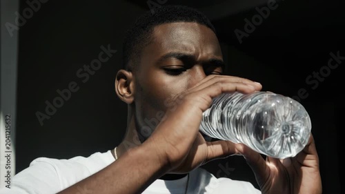 Slow motion static handheld close up of young man inspecting and drinking from plastic water bottle in indoor doorway with high contrast lighting