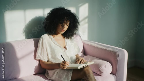 Static slow motion close up of woman writing in journal on pink sofa in sunlit living room interior afternoon